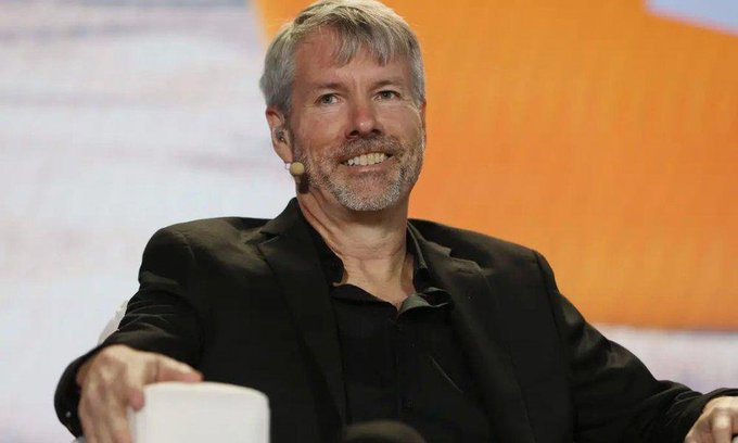 A photograph of Michael Saylor, executive chairman of Strategy (formerly MicroStrategy), smiling and speaking on stage while wearing a black blazer over a dark shirt.