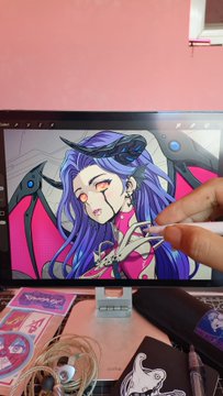 A hand drawing a pink anime-style female character with purple hair and horns on a digital tablet, with traditional ink and brushes visible on the desk below.
