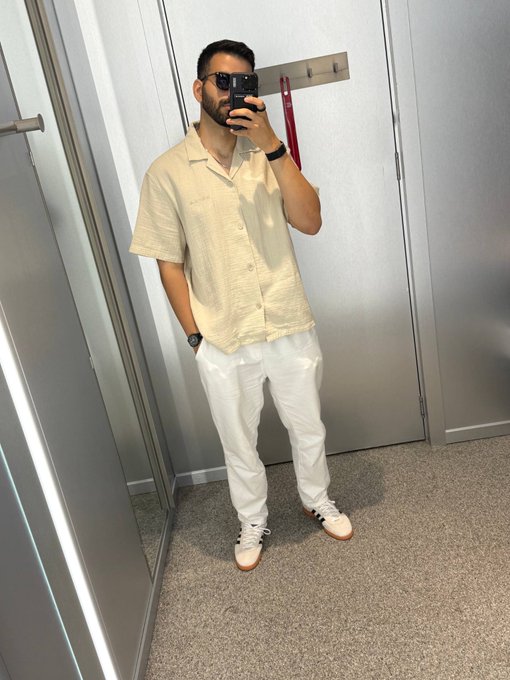A man wearing sunglasses and a light-colored button-down shirt paired with white trousers takes a mirror selfie in a fitting room, with the text "GM CT happy Wednesday" displayed.