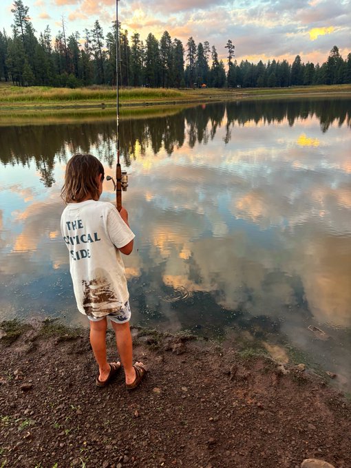 A child wearing a shirt reading 'THE MYSTICAL SIDE' stands barefoot at the edge of a calm lake holding a fishing rod, with pine trees and a sunset reflecting on the water.
