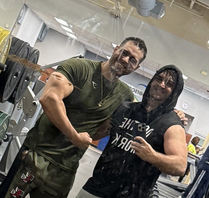A gym selfie of two men, with the man on the right taking the photo while flexing, their faces obscured by digital distortion effects.