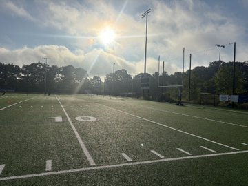 A sunlit American football field with yard markers, surrounded by trees and tall light posts under a partly cloudy sky.