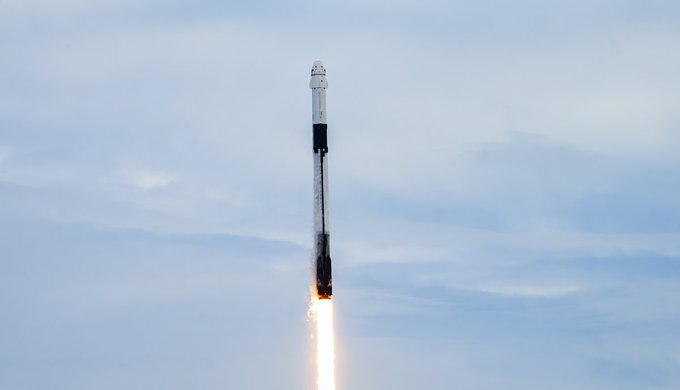 A SpaceX rocket ascends through a partly cloudy sky, its engines firing with a bright orange flame and a trail of exhaust smoke.