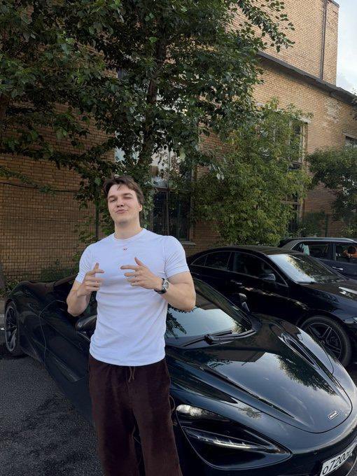 A young man in a white t-shirt and brown pants stands in front of a black McLaren sports car parked near a brick building, making hand gestures toward the camera.