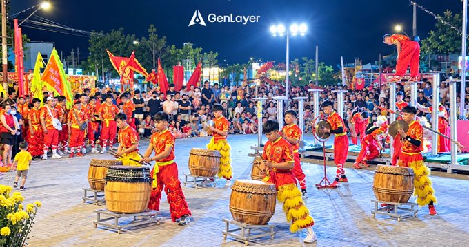 A vibrant nighttime cultural performance featuring drummers in red and yellow traditional attire performing with large barrels, with the 'GenLayer' logo displayed at the top.
