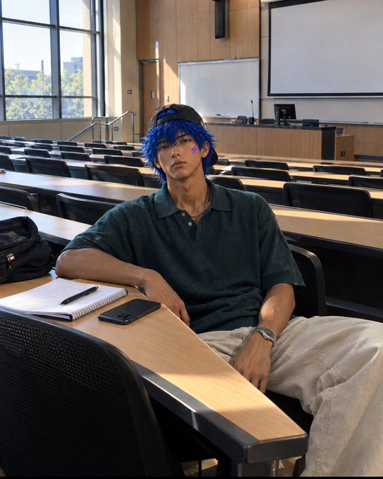 A person with bright blue hair wearing a black cap and dark polo shirt sits at a wooden desk in a lecture hall, with a notebook and phone placed in front of them.