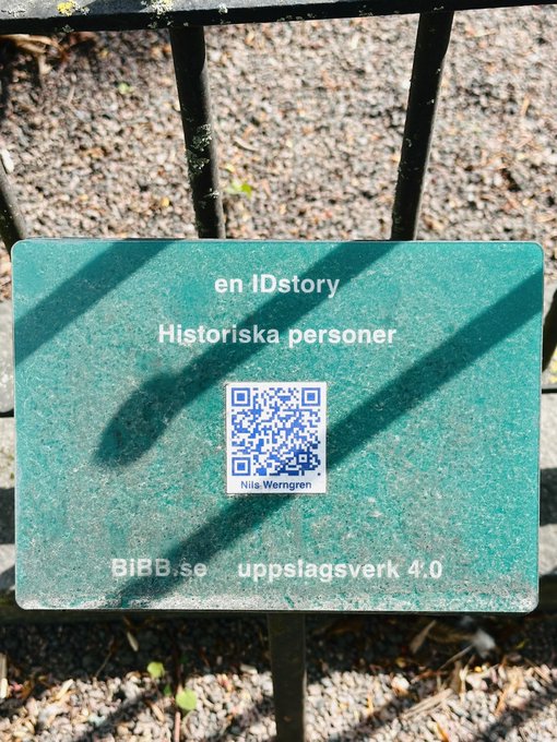 A teal informational sign mounted on a black metal fence, reading 'en IDstory', 'Historiska personer', and 'Nils.Werngren' above a QR code, with 'BiBB.se' and 'uppslagsverk 4.0' at the bottom.