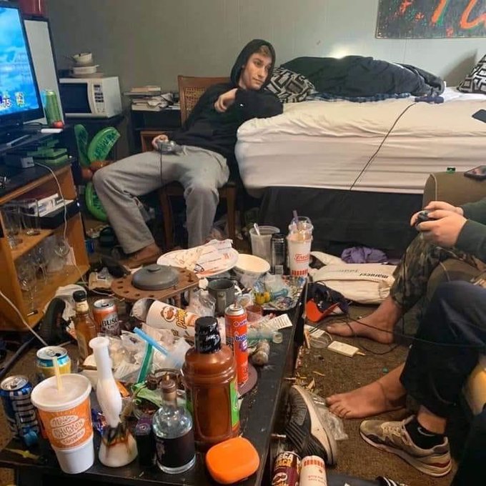 A messy dorm room scene with a person in a black hoodie sitting on a chair playing video games amid cluttered coffee cups, cans, and trash on a coffee table.
