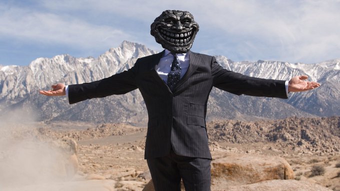 A figure in a black pinstripe suit stands with arms outstretched in a desert landscape, wearing a grinning troll face mask with mountainous terrain in the background.
