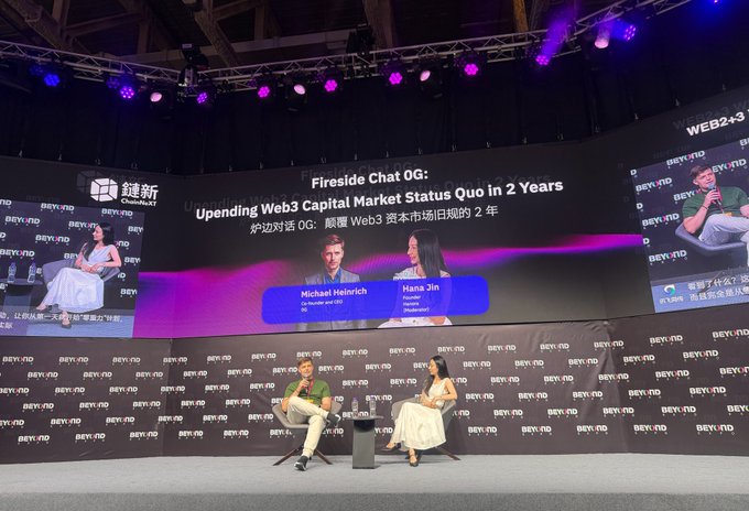 The image shows a stage with two people sitting behind microphones, likely participating in a fireside chat or panel discussion. The backdrop features a large screen displaying text related to the topic of the conversation. The speakers are positioned at different angles on the stage, suggesting an interactive and engaging event.