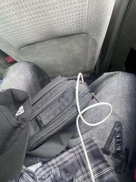A first-person view from an airplane seat showing a passenger's legs, a plaid blanket, a backpack, and a white charging cable on their lap.