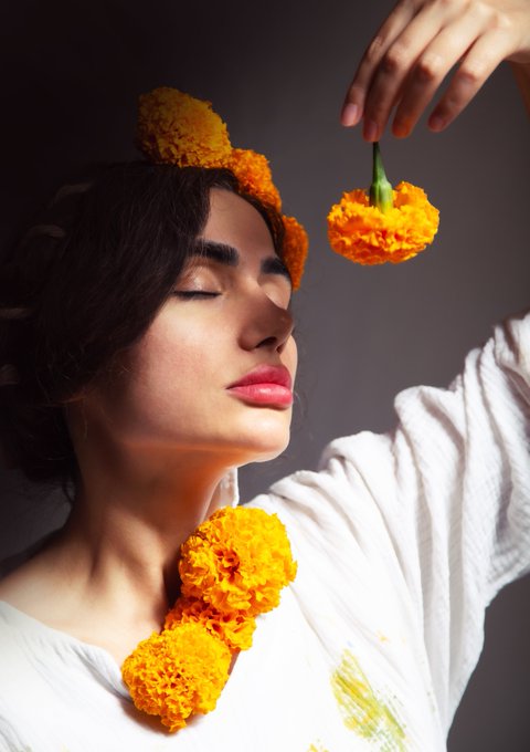 A woman with closed eyes wears orange marigold flowers in her hair and as a garland, holding another flower by its stem against a dark background.