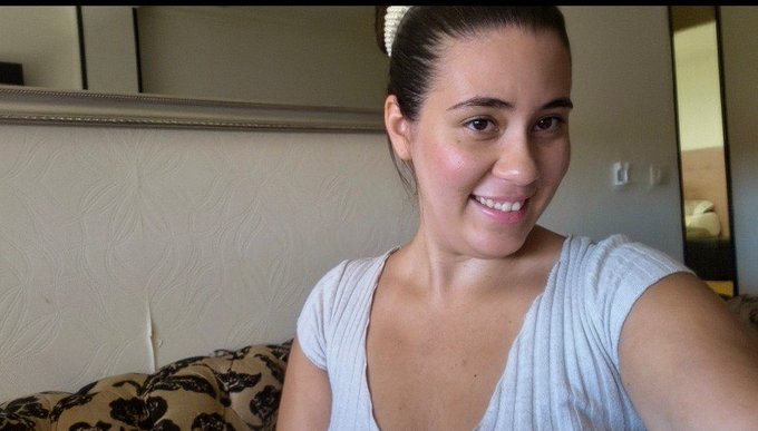 A smiling woman with dark hair pulled back takes a selfie, wearing a light gray top in what appears to be a hotel room.