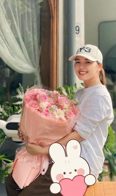 A smiling young woman wearing a white 'Y-3' cap and white long-sleeve shirt holds a large bouquet of pink roses wrapped in pink paper, with a cartoon rabbit sticker overlaid on the image.