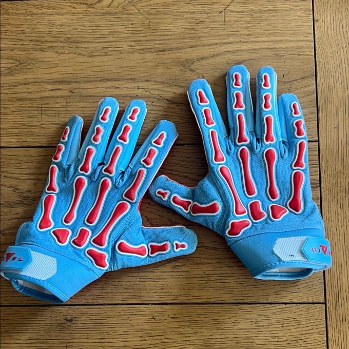 A pair of blue REV'IT! motorcycle/sports gloves with red and white skeleton bone graphics on the back of the hand, displayed on a wooden surface. The "REV'IT!" brand logo is visible on the wrist strap of each glove.