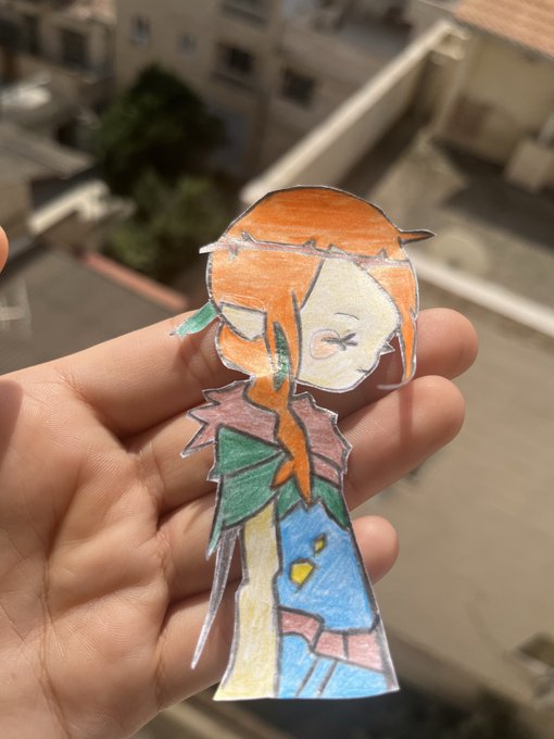 A hand holds a hand-drawn paper cutout of a character with orange hair and colorful clothing, photographed against an outdoor balcony background.