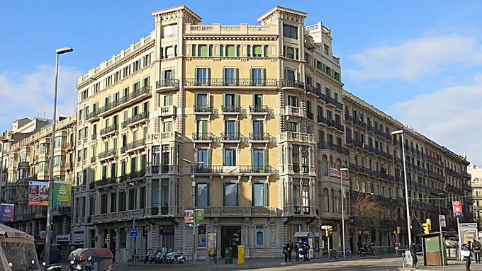 és-España, a historic yellow building with white accents and balconies, stands tall against the backdrop of a clear blue sky and cityscape.