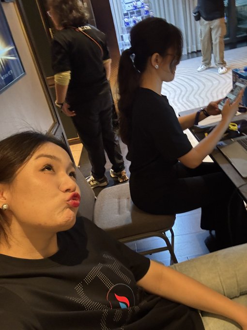 Two women are in a lounge setting; one in the foreground makes a playful pouting face at the camera, while another sits behind her looking at her phone.