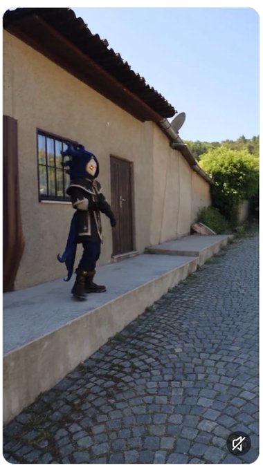 An animated character in blue and gold armor with a dragon-like hood stands on a ledge along a tan building with a cobblestone path.