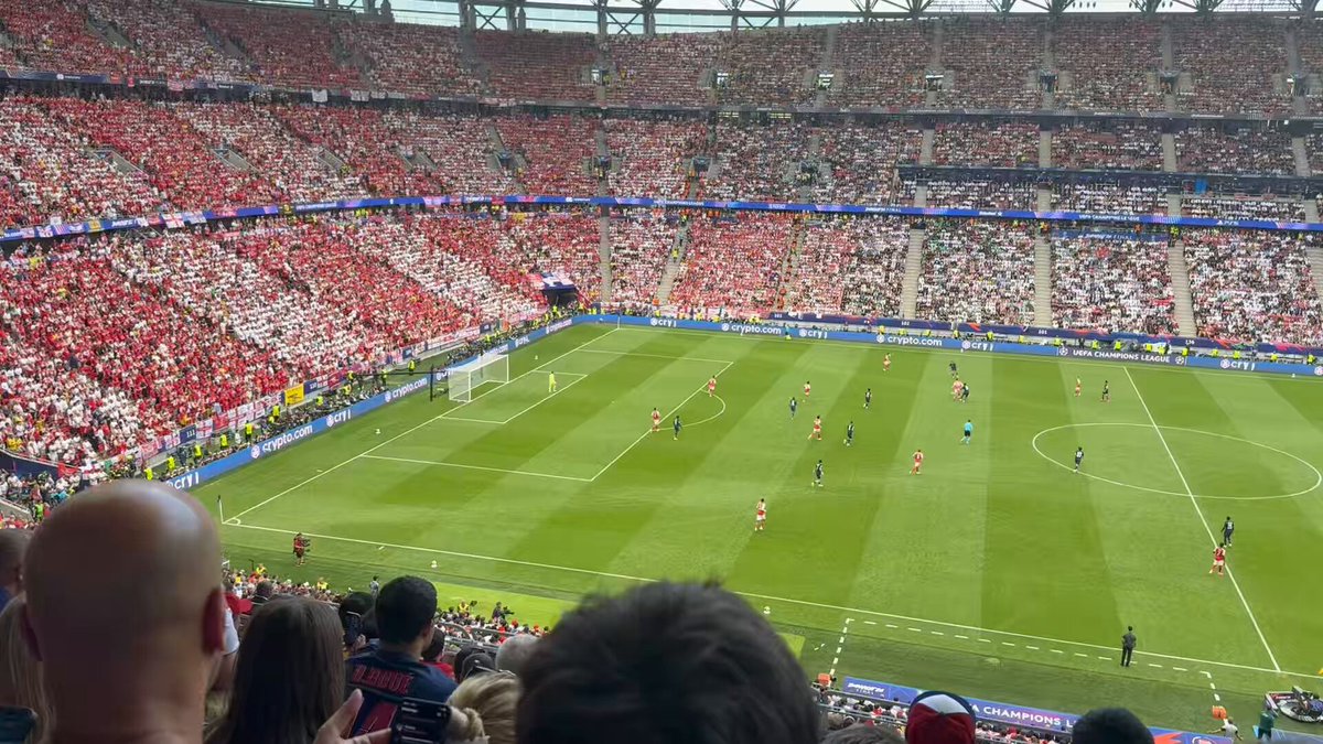 The image shows a large soccer stadium filled with spectators, where two teams of players are engaged in an intense match. The crowd is seated in rows of chairs, watching the game intently from their seats. The atmosphere appears lively as fans cheer for their favorite team and enjoy the thrilling competition unfolding on the field.