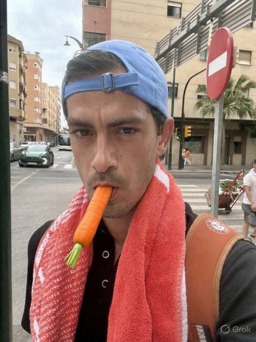 A selfie of a young man wearing a blue backwards cap and a red towel around his neck, holding a carrot in his mouth on a city street with a "Do Not Enter" sign visible.