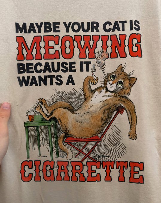 A graphic t-shirt design with the text 'MAYBE YOUR CAT IS MEOWING BECAUSE IT WANTS A CIGARETTE' above an illustration of a cat lounging in a chair, smoking a cigar with a drink on a side table.