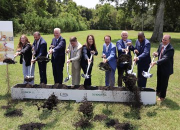 A group of people in formal attire participate in a groundbreaking ceremony, holding shovels with dirt over a wooden marker displaying logos for Google, FIS, and Deutsche.