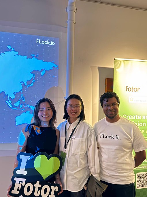 Three people pose together at a Fotor x FLock.io event, with a projection of a world map displaying the "FLock.io" logo behind them and a green "fotor" banner to the side. The woman on the left holds an "I ❤️ Fotor" sign, while the man on the right wears a t-shirt reading "FLock it.", reflecting the collaborative Video Agent launch event where FLock's Head of EMEA and Head of Institutional Development attended alongside the Fotor team.