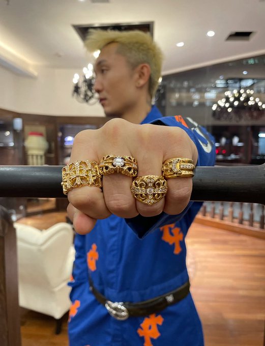 A man in a blue and orange outfit displaying four large, ornate gold rings with gemstones on his fingers.