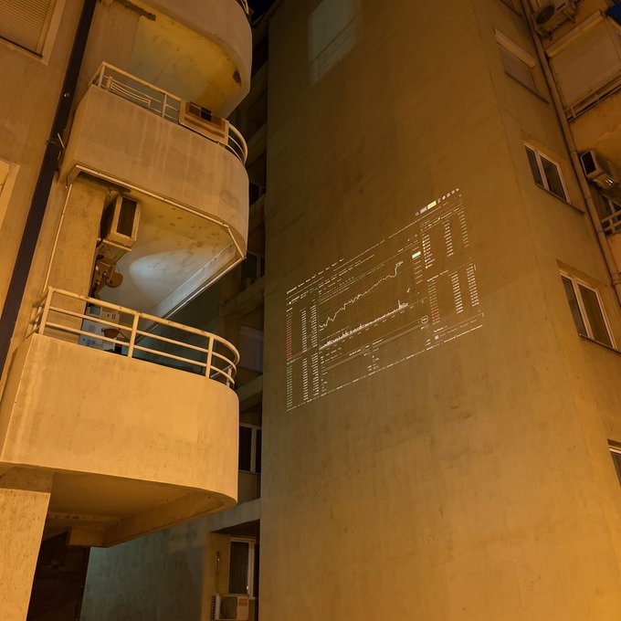 A nighttime projection of a candlestick stock chart (with "PEPE/USDT" and "Binance" visible) is cast onto the side of a residential apartment building with curved balconies, humorously illustrating a trader's obsession with crypto markets even at home.