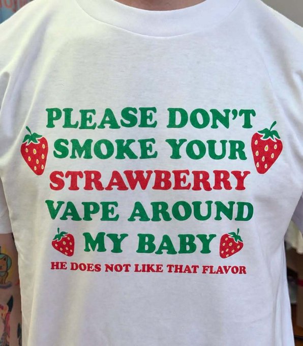 A white t-shirt with a festive design reading 'PLEASE DON'T SMOKE YOUR STRAWBERRY VAPE AROUND MY BABY HE DOES NOT LIKE THAT FLAVOR,' decorated with red strawberries.