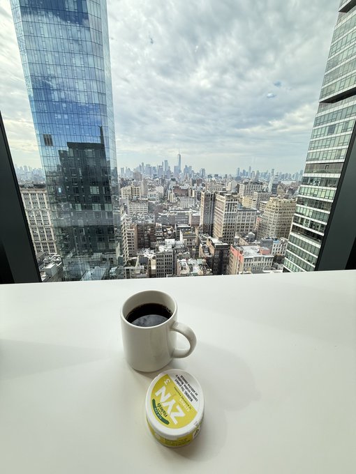 A cup of coffee sits beside a "TRU-RAN" cream pod on a high-rise windowsill, overlooking the expansive city skyline of New York, including the Freedom Tower—a fitting visual for "bringing the world onchain," symbolizing the city's global reach from above.