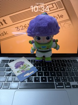A small Buzz Lightyear figurine with purple hair sits on a laptop keyboard next to a matching card, with a notebook page displayed on the screen behind it.