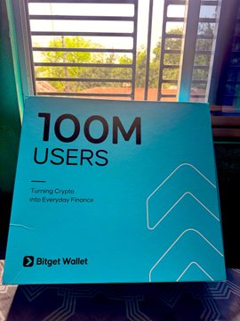A Bitget Wallet promotional poster celebrating '100M USERS - Turning Crypto into Everyday Finance' displayed near a window.