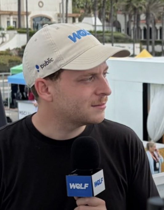 A man wearing a beige "Public" cap and black shirt is being interviewed with a microphone branded "WOLF" and "WOLF" visible on his hat, displaying a concerned side-glance that humorously matches the post's caption about losing money on a prediction market trade if interest rates are raised.