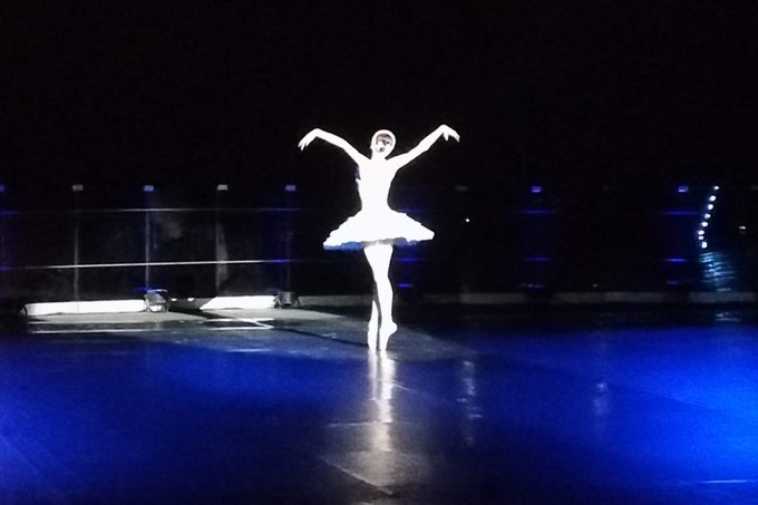 A ballerina in a white tutu stands on pointe in a spotlight, illuminated against a dark blue stage. The image, titled "Dancing through the shadows - On tiptoe," captures the ethereal beauty of ballet performance.