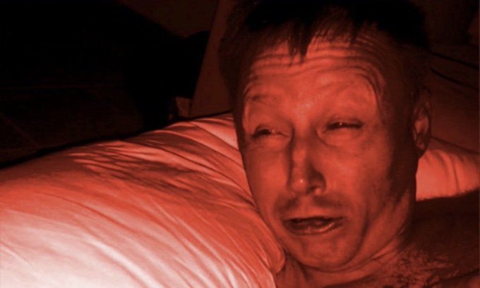 A grainy, red-tinted photo of a man lying in bed against white pillows, appearing distressed with his eyes closed.
