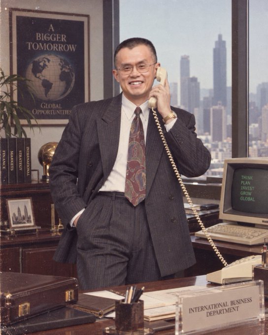 A businessman in a pinstripe suit and paisley tie smiles while holding a vintage telephone in an office with a "International Business Department" sign, a "A Bigger Tomorrow" globe poster, and a monitor displaying "Think Plan Invest Grow Global." The retro corporate setting visually complements the post's reference to the "1998 digital cash research plan by Wei Dai," evoking the era when foundational cryptography ideas like b-money were first proposed on early mailing lists before Bitcoin's eventual creation.