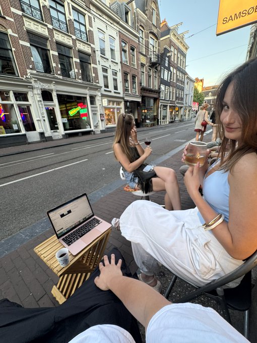 Two women enjoy coffee and work on a laptop at a small wooden table on a sunlit Amsterdam street, capturing the relaxed outdoor work scene mentioned in the post about moving the "office" outside with @ginie_exe and @PotBot_sol.