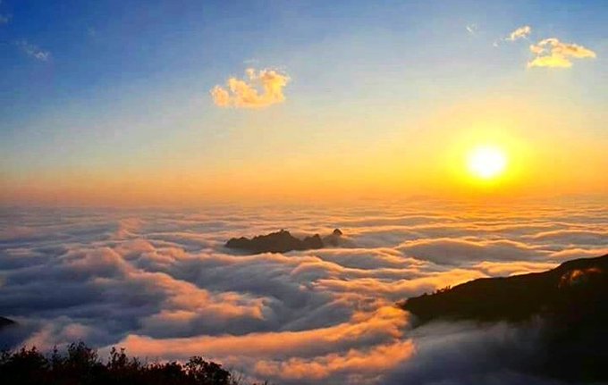 A scenic view of a bright sun setting over a sea of clouds with mountain peaks visible in the distance.