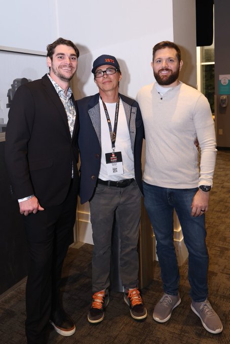 Three men pose together at an indoor event, with the central figure wearing a navy "1971" cap, glasses, a blazer over a white tee, and a lanyard with what appears to be a "Film" badge. The caption honors RJ Mitte (right), noting a recent conversation about Bitcoin, while the middle man is likely Adam Simecka, who introduced them.