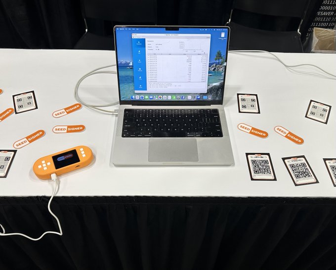 A laptop on a table at the @MWBTCSummit displays SeedSigner setup screens, accompanied by an orange SeedSigner hardware device and QR code informational cards arranged around it, showcasing the Bitcoin signing tool available for demos at the exhibition hall table.