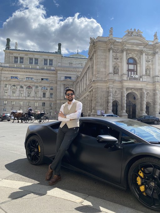 A man in a white shirt, cream vest, and sunglasses leans confidently against a matte black Lamborghini sports car in a European plaza, with an ornate baroque building and horse-drawn carriages in the background.
