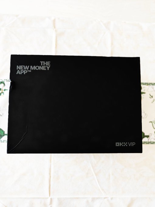 A black envelope with 'THE NEW MONEY APP™' printed on it, resting on a white fabric surface.