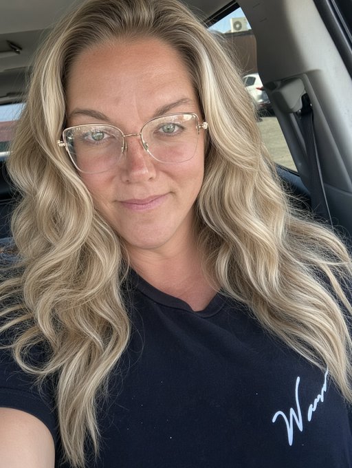 A blonde woman wearing clear-framed glasses and a dark t-shirt with white script text takes a selfie while sitting inside a car.