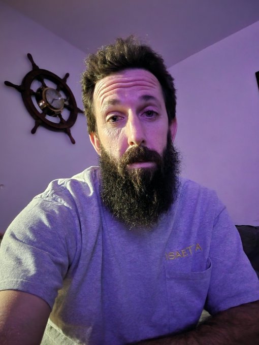 A bearded man wearing a grey t-shirt sits indoors beneath a decorative ship's wheel mounted on a purple-lit wall.