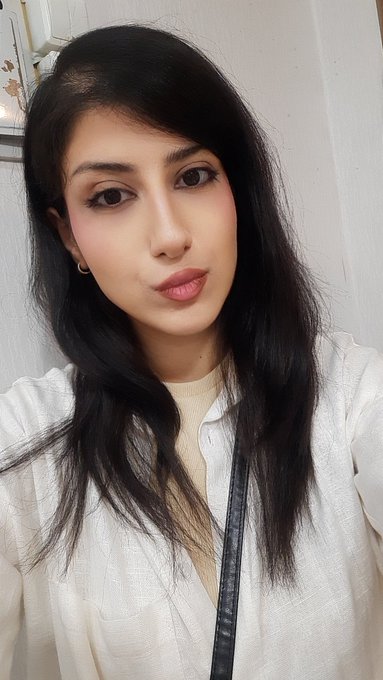 A close-up portrait selfie of a young woman with long dark hair, wearing a white shirt over a beige top, looking directly at the camera.