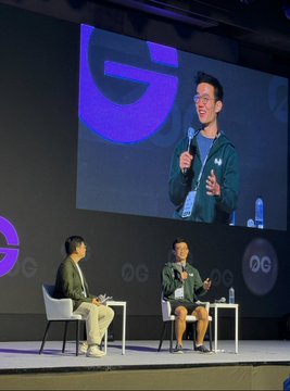 A conference stage shows two people seated while a third person is displayed on a large screen above them speaking into a microphone.