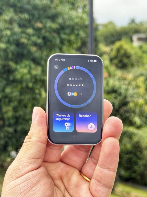A hand holds a small foldable smartphone outdoors, displaying a circular widget with “Chaves de segurança” and “Receber” buttons.