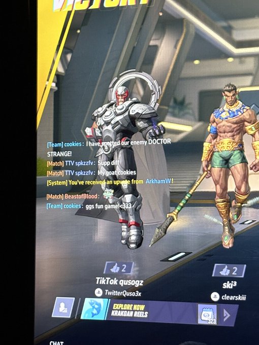 A gamer is showing off a post-victory screen from a video game featuring two characters—one robotic armored figure and one muscular warrior with a spear. The chat overlay shows messages from teammates like "cookies" and "TTV spkzzfv," with an "EXPLORE NOW" button for KRANKAN REELS, and the player appears pleased about being shouted out by a streamer named "Luna."